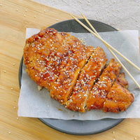 A plate of sliced chicken tenders in golden sweet sauce, sprinkled with sesame seeds. Place the chicken on a piece of parchment paper on the plate. There are two wooden skewers on the side, reminiscent of Taiwanese night market food. The plate sits on a wooden surface.