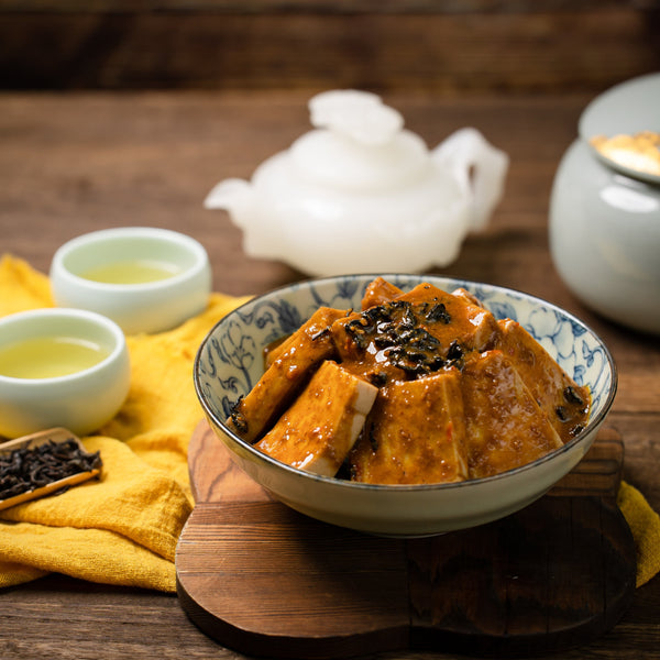 A bowl of Braised Tofu in Signature Sauce, filled with tofu chunks, red chili sauce, with black beans, on a wooden stand, with tea and a white teapot in the background.