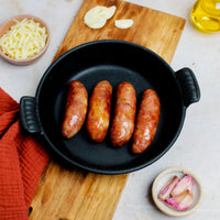 Four grilled Taiwan cheese sausages sit in a black cast iron skillet, with a wooden cutting board, grated cheese, garlic cloves, oil, and chopped onions on a nearby marble countertop.