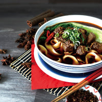 A bowl of Taiwan Beef Noodle Soup with scallions and cabbage, surrounded by cinnamon sticks, star anise and pepper on a wooden table.