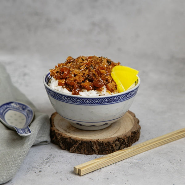 On a wooden coaster sits a bowl of rice topped with delicious Taiwanese pork jerky with sliced kimchi. A pair of chopsticks and an oriental spoon rest on a napkin nearby. In the background is a plain, textured surface.