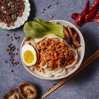 A bowl of noodles in a minced pork soup topped with a poached egg, cabbage and mushrooms. Around the bowl are dried chili peppers, black pepper, and a dish of mixed spices. The rich, spicy sauce adds flavor to this ready-to-eat meal. Chopsticks are placed next to the bowl.