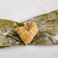 The light-colored surface holds a triangular glutinous rice dumpling wrapped in bamboo leaves. A visible chestnut is embedded in the top of the dumpling, and the bamboo leaves show a slight luster and green color, enhancing the delicious presentation.