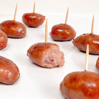Smoothly glazed small sausages, each skewered on a wooden toothpick, crispy on the outside and tender on the inside, are arranged on a white surface.