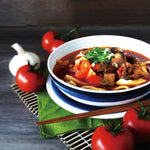 A bowl of slow-cooked beef noodle soup with tomato, green onions, surrounded by fresh tomatoes and garlic, with a pair of chopsticks on top of the bowl, placed on a dark wooden table.